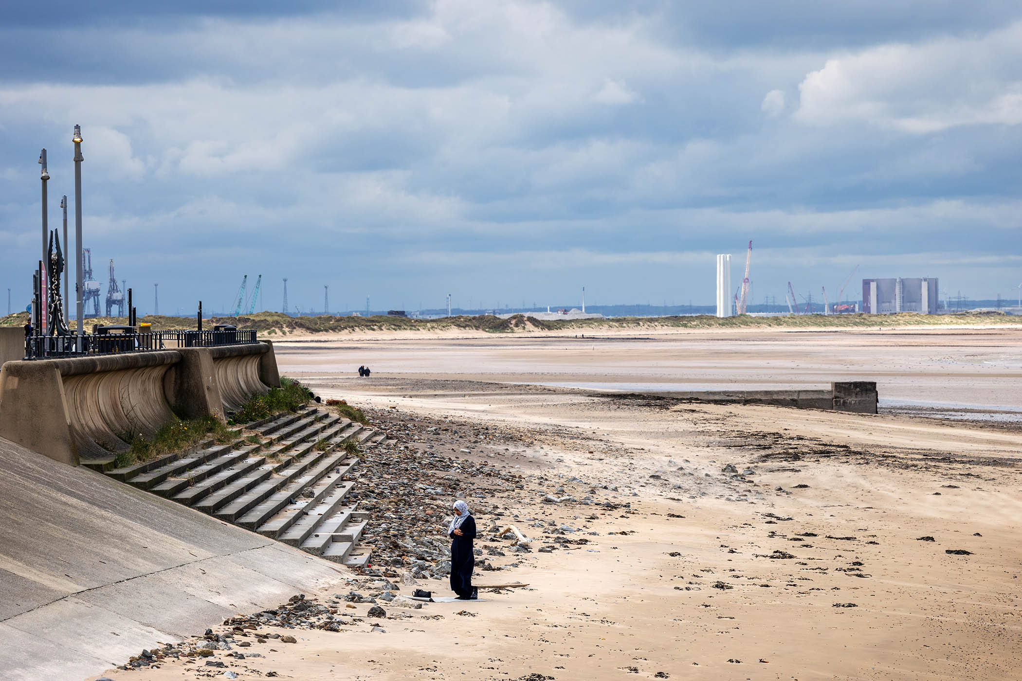 A woman takes midday prayers on Redcar beach with the former Corus steel works and Teesworks site in the background
